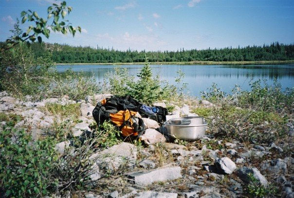 A black and orange backpack with a seive and sampling bowl by a lake surrounded by evergreen trees in summer.