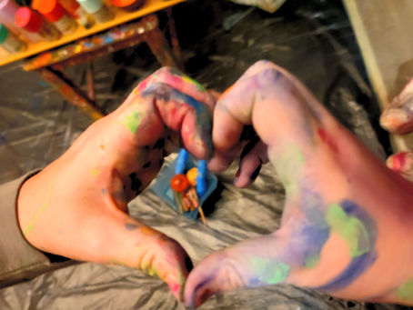 Paint-covered hands form a heart shape. Background shows colorful paint bottles on a table. Creative and playful mood.