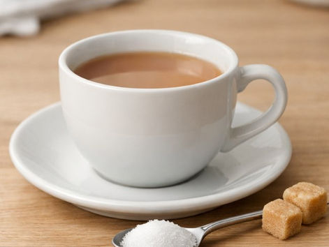 Cup of tea on a wooden table with a teaspoon of sugar and two sugar cubes beside it, in soft natural light