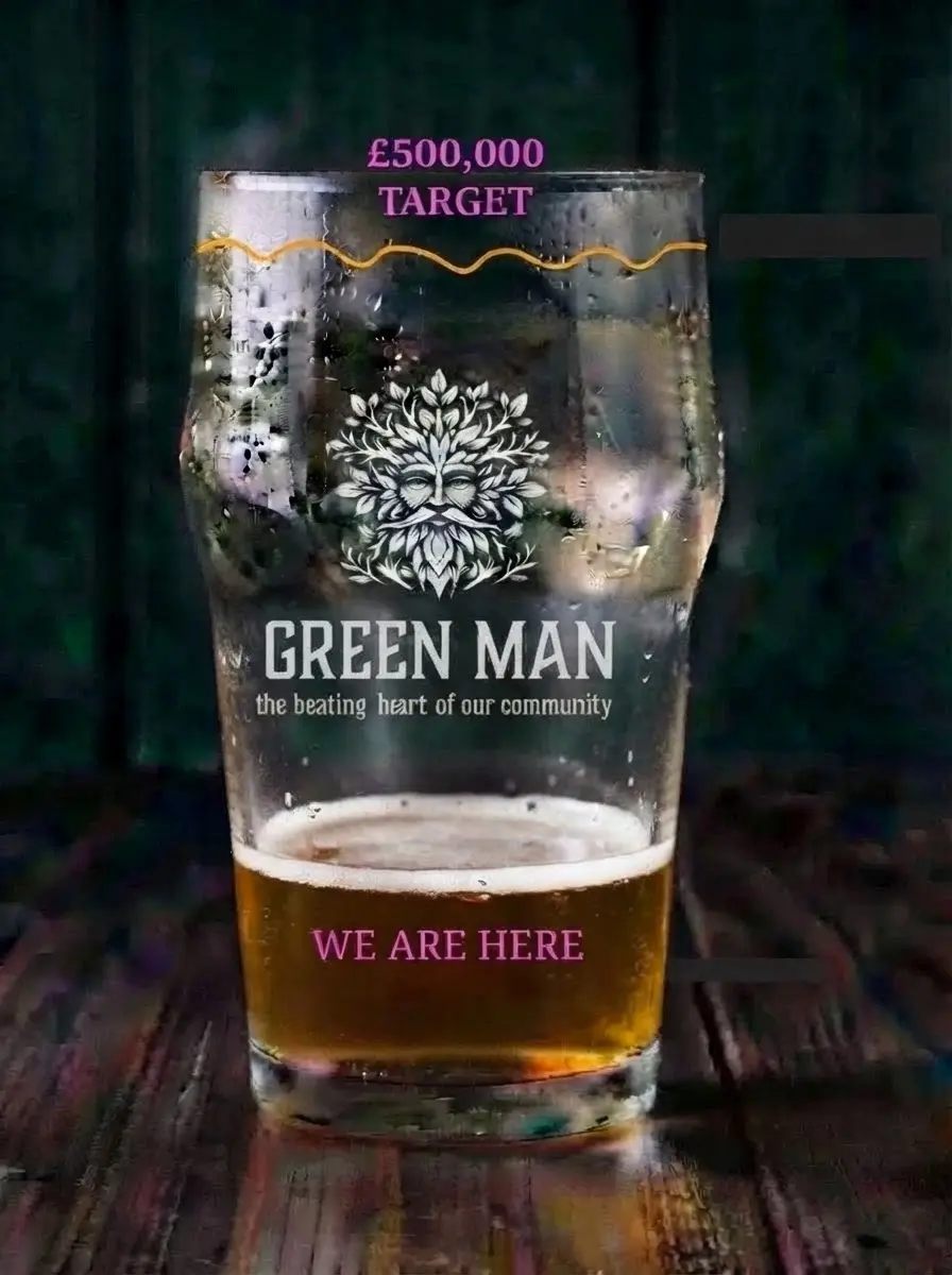 Studio photography of a pint glass filled with beer, featuring a 'Green Man' branding logo printed on the glass. The background is a dark, rustic wooden surface creating a cozy pub atmosphere.