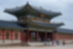 Historic Korean palace gate with ornate details; people in traditional clothing walk nearby, under a cloudy sky.