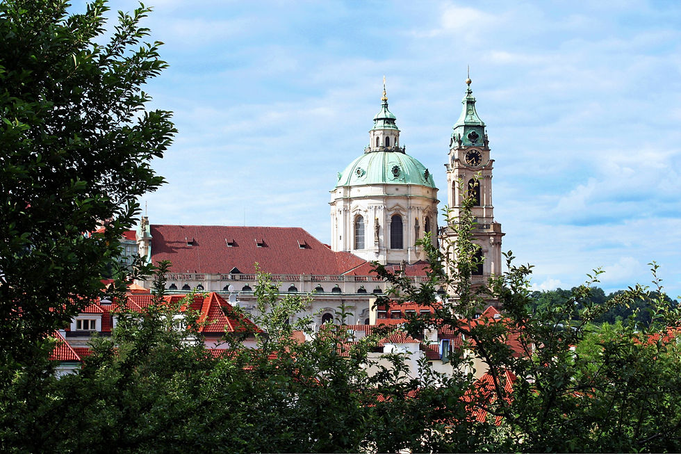 Historic church with green domes and clock tower against a blue sky. Red-roofed buildings and lush green trees in the foreground.