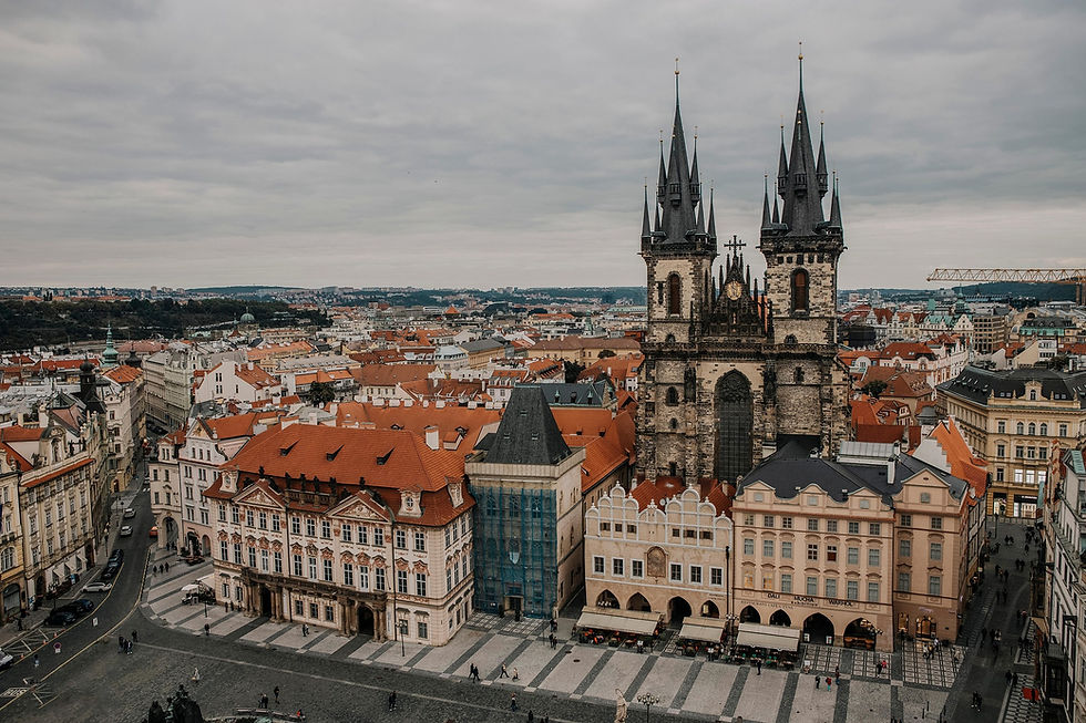 Aerial view of a historic city square with ornate buildings, red roofs, and a Gothic church. Overcast sky sets a somber mood.