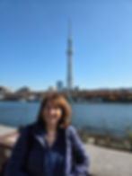 Woman in a blue jacket smiles by river with Tokyo Skytree in background. Clear blue sky, cityscape with autumn trees, and calm water.