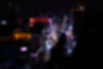 Night cityscape with vibrant neon lights on buildings lining a bustling street. Cars drive along wet pavement, with a dark urban backdrop.
