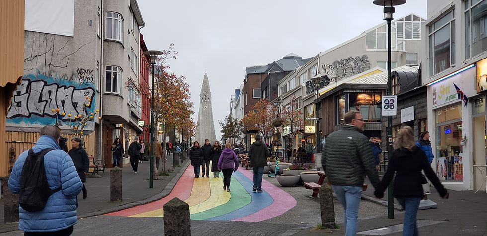 Pedestrians walk along a rainbow-painted street in a gray city center, with graffiti walls, shops, and a distant church spire.