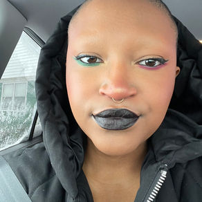 A young Black woman with brown skin and a shaved head and colorful makeup gazes at the camera. Black lipstick paints her full lips. A septum ring hangs under her nose. Under one eye, green eye liner has been drawn on. Dark pink is under the other eye. 