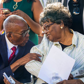 Seated in a wheelchair an elderly Black woman, Claudette Colvin, with brown skin smiles while leaning toward an elderly Black man in a blue suit, Fred Gray. 