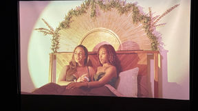 In a bedroom filled with soft natural lighting, two Black women, one light-skinned, one dark-skinned, sit close in bed against a wooden headboard with golden metal slats that radiate outward like a sunrise with fronds of green plants on top.