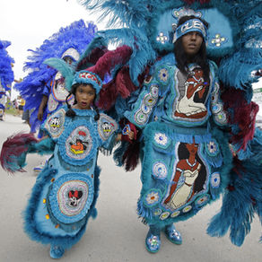 A little Black girl wearing a sky blue costume: a headband with a blue feather, a chest plate with a beaded cartoon Goofy in the center and Minnie Mouse on the skirt and blue flats, holds hands with her mother in a long-sleeved costume of the same sky blue color, except hers has a woman that resembles Pocahontas on the chest and skirt. 