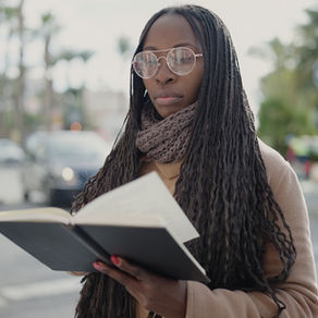A Black woman with dark-skin and long braids reads a hardcover book while standing on the street 