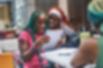 Two young Black women one with teal braids, one with orange-red braids, huddle together and smile down at a notebook.