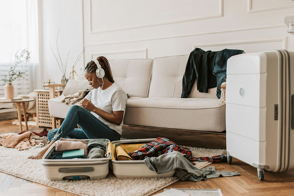 A young Black woman with braids sits on the floor with an open suitcase next to her and a second, closed suitcase a short distance away.