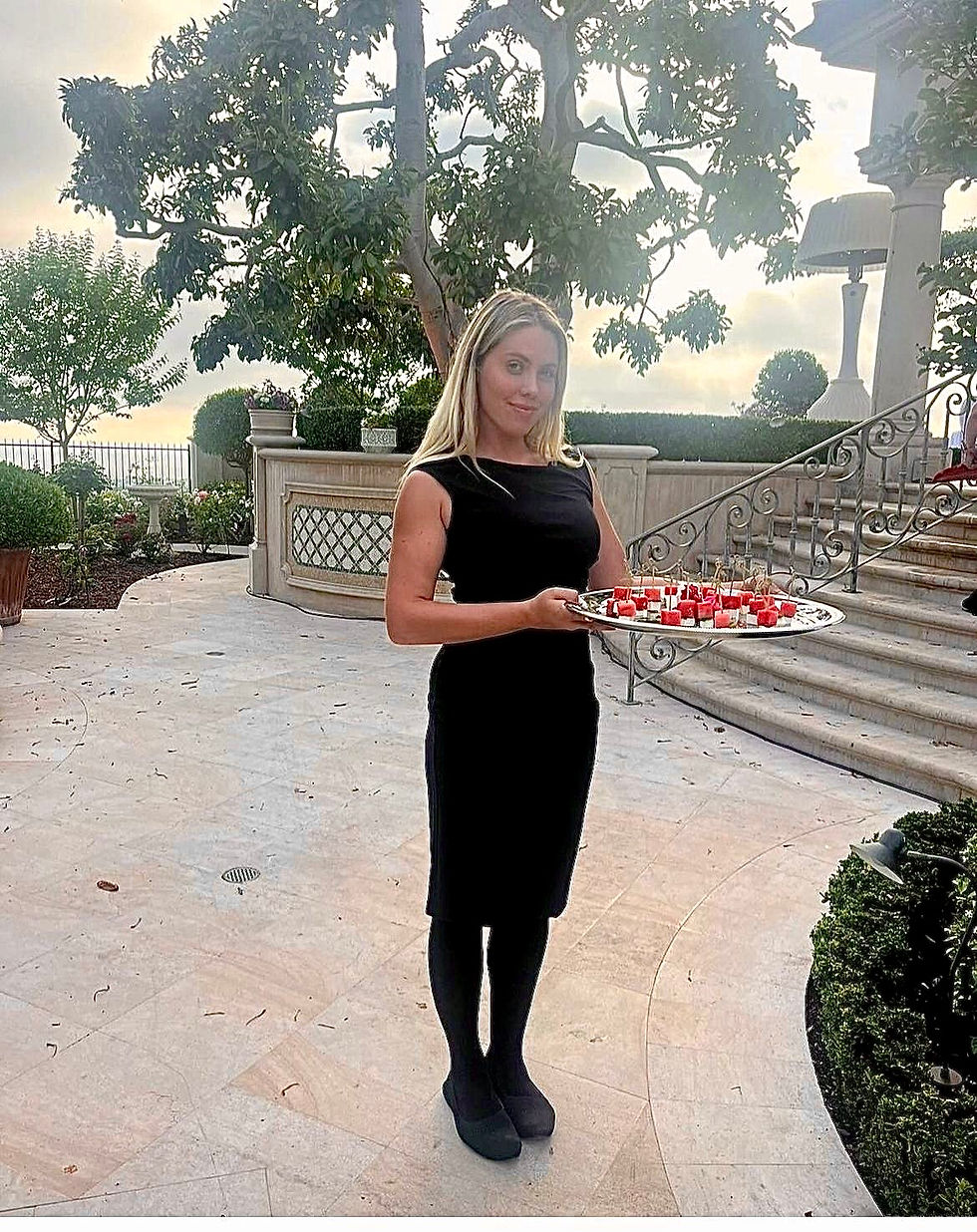 Woman in black dress holds a tray of appetizers outdoors. Stone patio, trees in background, elegant setting, calm evening ambiance.