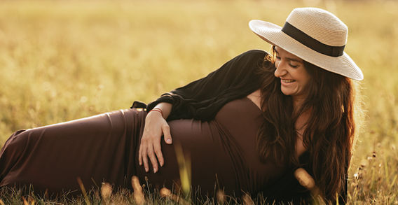 Žena leží na louce, těhotenská fotografie, usmívající se, s kloboukem a slunečným dnem.