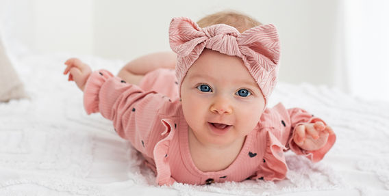 Roztomilé dítě leží na bílé dece, usmívající se, s mašlí na hlavě.