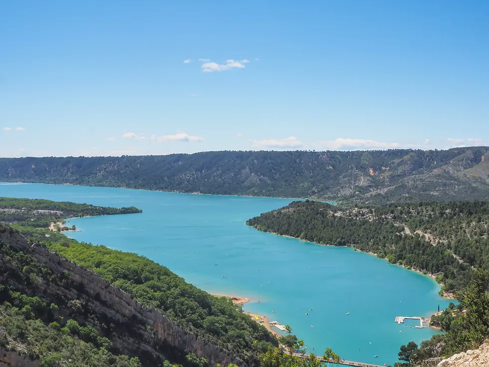 A long view of Lac de Sainte-Croix taken from a nearby road overlooking the lake. The banks of the river are largely covered in trees, other than for a small beach on the nearside of the lake