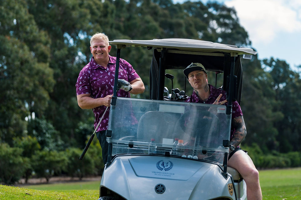 Two men, Ben Hannant and Chris McQueen, in FOGS Maroon Golf polos, one standing outside and the other seated in a golf cart, on a golf course with trees in the background.