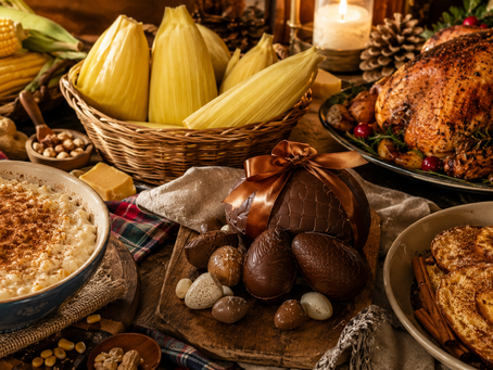 Uma grande mesa horizontal dividida de forma orgânica pelas estações e celebrações culturais. Em destaque, uma canjica fumegante e uma pamonha remetendo às festas juninas, ovos de chocolate sofisticados representando a Páscoa e uma mesa natalina elegante com peru dourado e rabanadas. Todos os elementos conectados visualmente por uma mesma narrativa de tradição, memória e pertencimento.