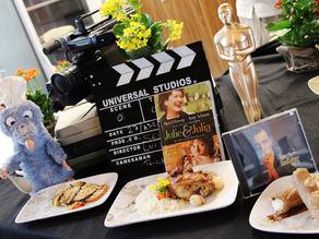 um chef preparando um prato inspirado em um filme, com uma tela exibindo a cena ao fundo.