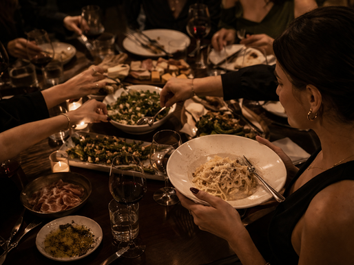 Imagem editorial de uma mesa compartilhada com vários pratos ao centro, enquanto uma pessoa segura seu prato próximo ao corpo, em contraste com mãos que se estendem para os outros — luz quente e atmosfera sofisticada