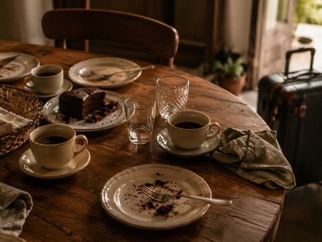 Uma mesa parcialmente ocupada após uma refeição de despedida. Xícaras de café ainda quentes, pratos com pequenos vestígios de sobremesa, guardanapos dobrados às pressas e uma cadeira vazia em destaque. Ao fundo, uma mala próxima à porta sugere partida iminente. A cena deve transmitir afeto, silêncio e a delicadeza dos encerramentos.