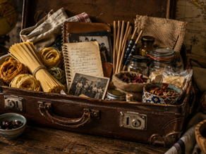 Uma mala de viagem antiga aberta sobre uma mesa de madeira. Em vez de roupas, ela está preenchida por elementos gastronômicos de diferentes culturas: massas italianas artesanais, hashis japoneses, especiarias árabes, pão artesanal, cadernos de receitas envelhecidos e fotografias de família. Ao fundo, um mapa antigo desfocado sugere deslocamento e passagem do tempo.