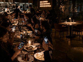 Imagem editorial de um restaurante contemporâneo lotado, com pessoas fotografando pratos e brindando, luz baixa e atmosfera sofisticada, em contraste com uma mesa vazia desfocada ao fundo