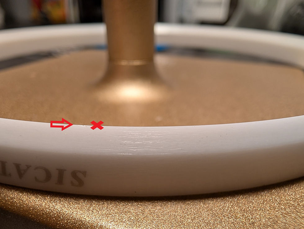 Nahaufnahme eines weißen Keramik Rakelrings auf goldener Oberfläche. Roter Pfeil und X markieren einen Makel. Unscharfer Hintergrund. Text "SICAT" sichtbar.