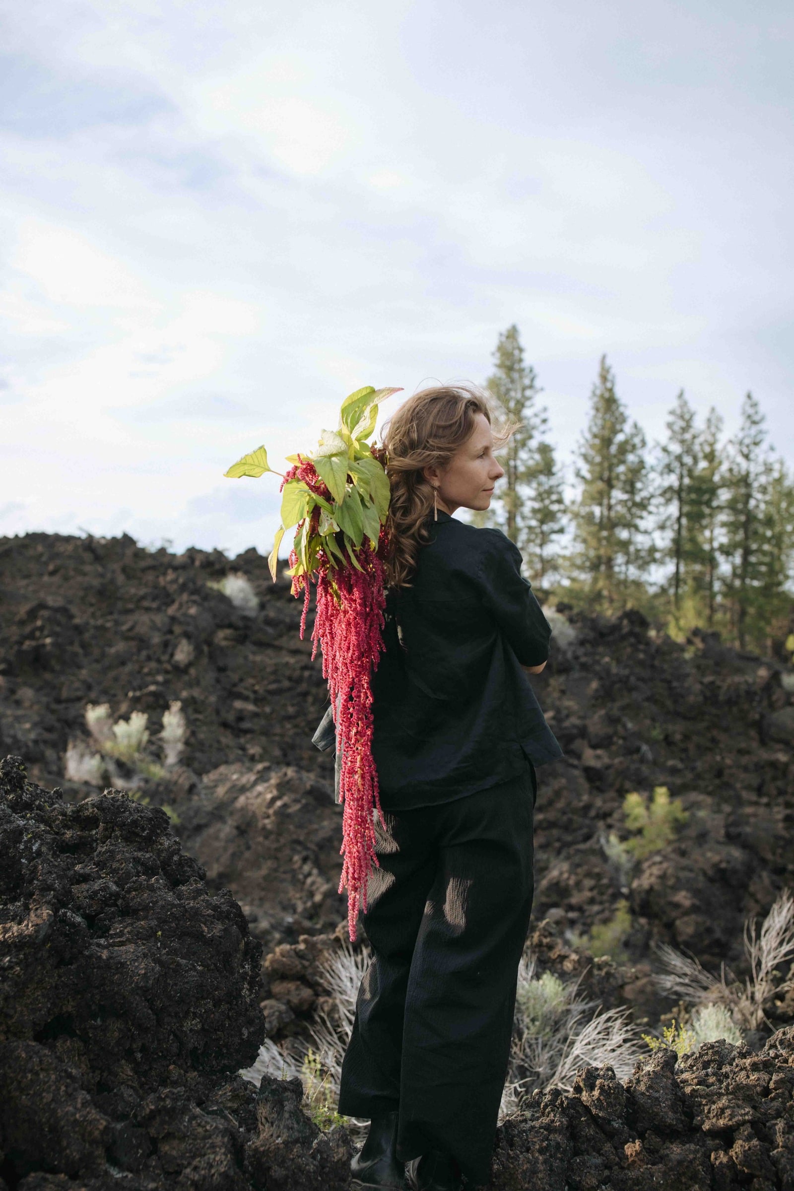 Perso in black clothing standing on volcanic rock holding a trailing red amaranth bouquet over her shoulder