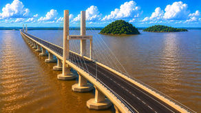 Aerial view of Batang Lupar 1 Bridge in Sarawak stretching across wide brown river waters under a bright blue sky with lush green islands in the background.