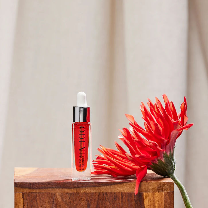 Sérum Oden rouge vif posé sur un socle bois, accompagné d’une fleur graphique – visuel de campagne.