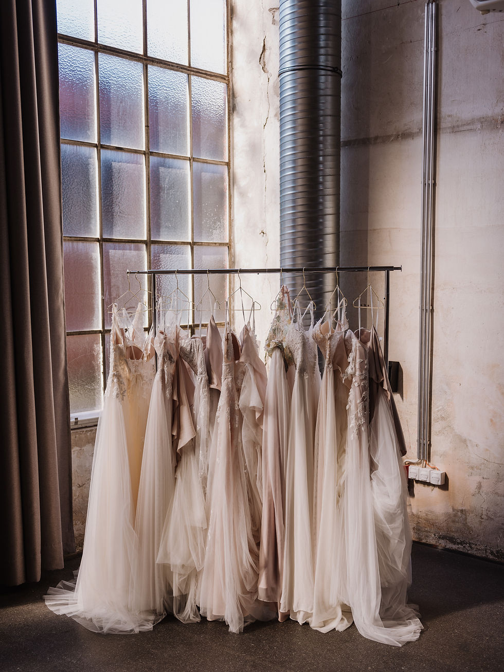 Several flowy ivory and white wedding dresses hanging from a rack in front of a large window