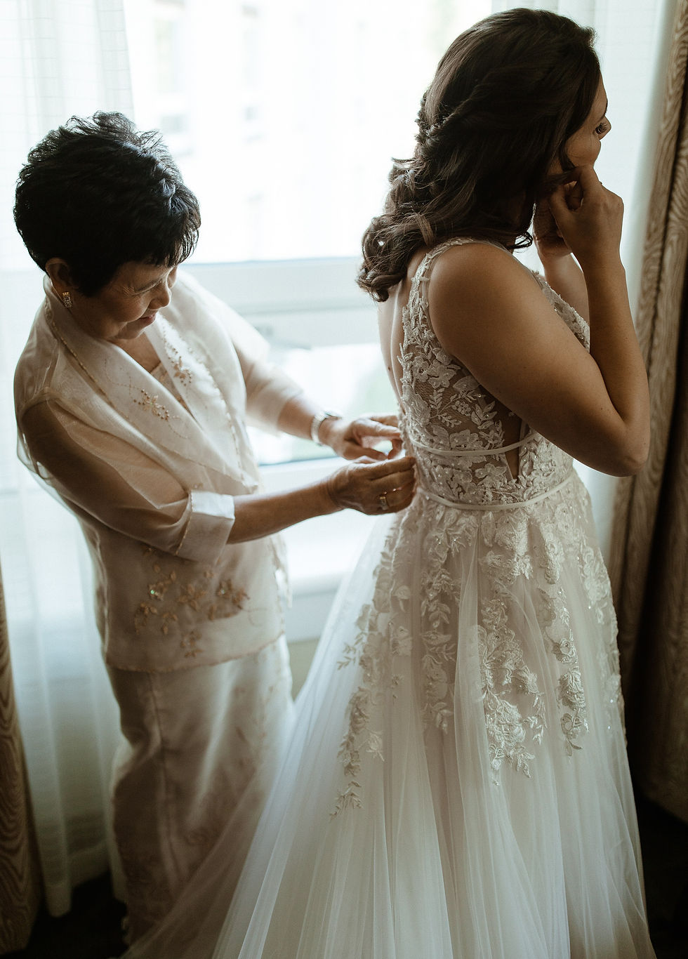 Bride putting on earrings while mom buttons up the back of a gorgeous lace wedding gown.