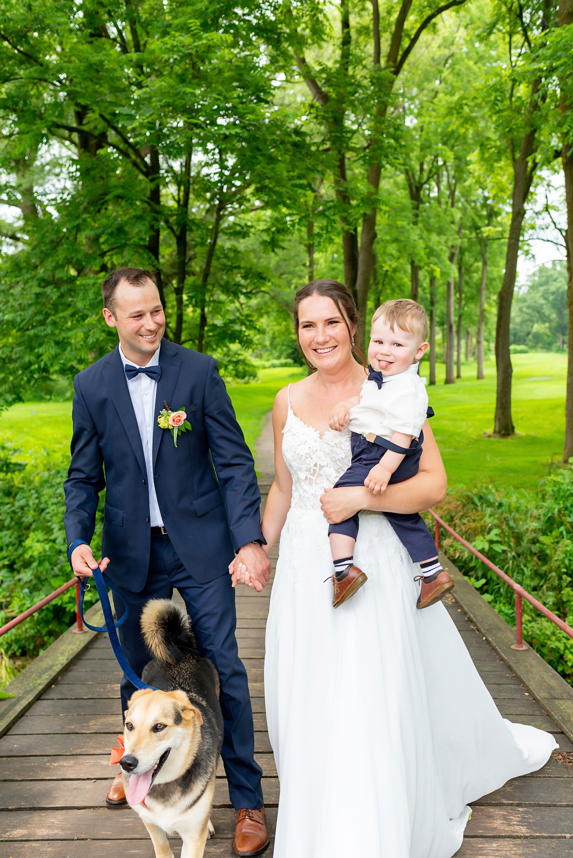 A family of three, parents in their wedding attire, walking over a bridge with a dog