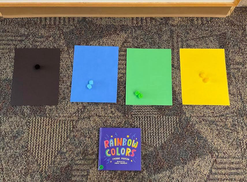 Colored sheets with matching pom-poms placed on each sheet—black, blue, green, yellow. Below them is a book titled "RAINBOW COLORS."