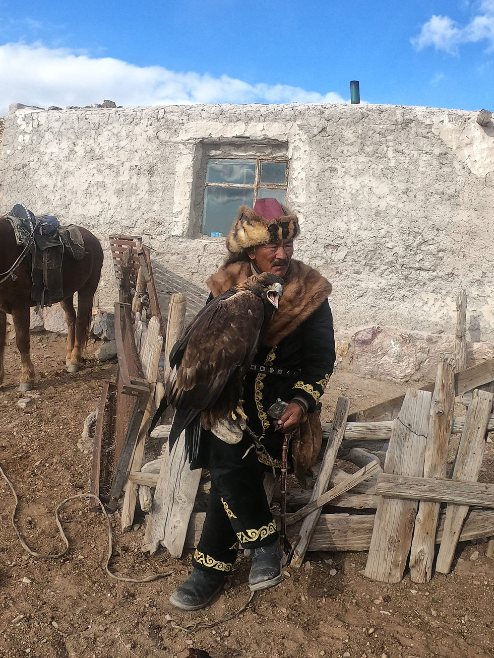 Kazakh eagle hunter in a fur hat and embroidered black coat holding a golden eagle on his arm beside a whitewashed house, a saddled horse to the left