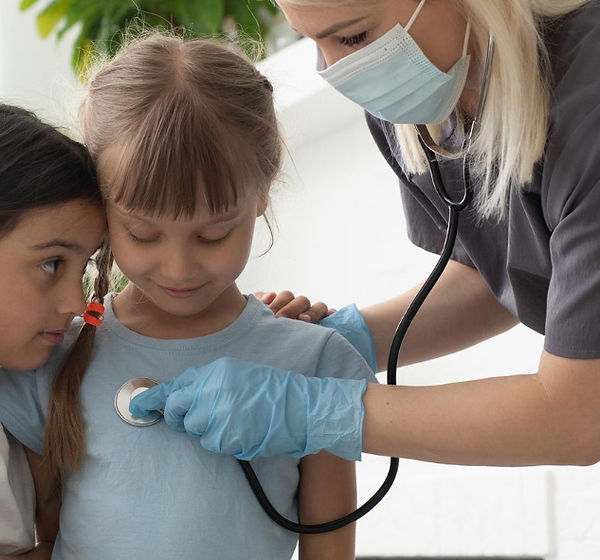 A healthcare worker in gray scrubs and mask checks a child's heartbeat with a stethoscope. Another child watches closely. Bri
