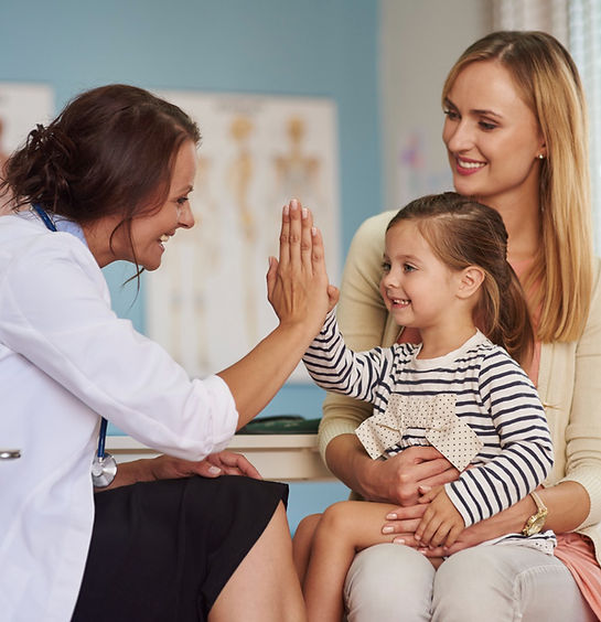 Doctor high-fives smiling child on mother's lap in a bright clinic. Blue walls, anatomical posters in the background. Warm, f