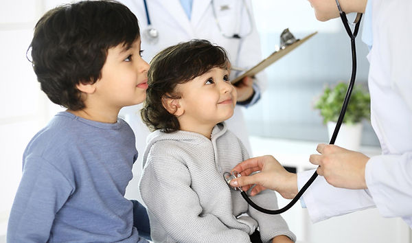 Two children smiling during a check-up as a doctor listens with a stethoscope. Background includes a clipboard and potted