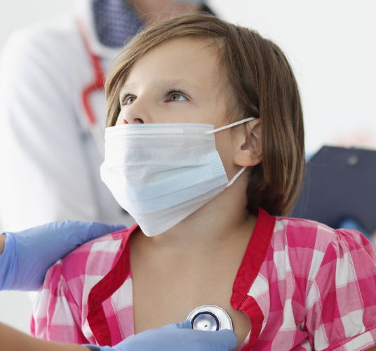 Child in pink checkered shirt and mask, being examined with stethoscope by medical staff in blue uniform and gloves, holding