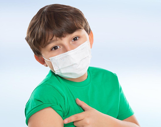 Child in green shirt wearing a mask points to a bandage on their arm, against a light blue background, indicating a recent va