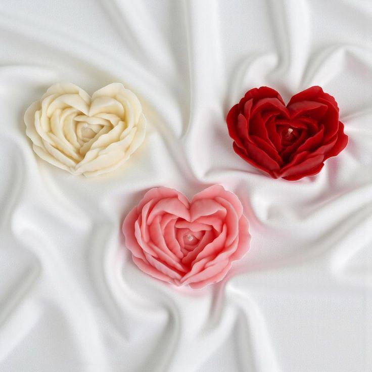 Three handmade rose heart candles in ivory, red and pink arranged on white satin fabric
