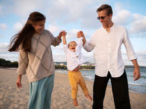 Parents walking on the beach with their child, each holding onto one of the child's hands and lifting him up
