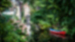 A red canoe sitting on a river lined in trees and that travels under an medieval bricked bridge with a parked motorcycle.