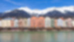 A row of pastel-colored houses in Austria with a blue sky, snowcapped mountain behind and a flowing river in front.