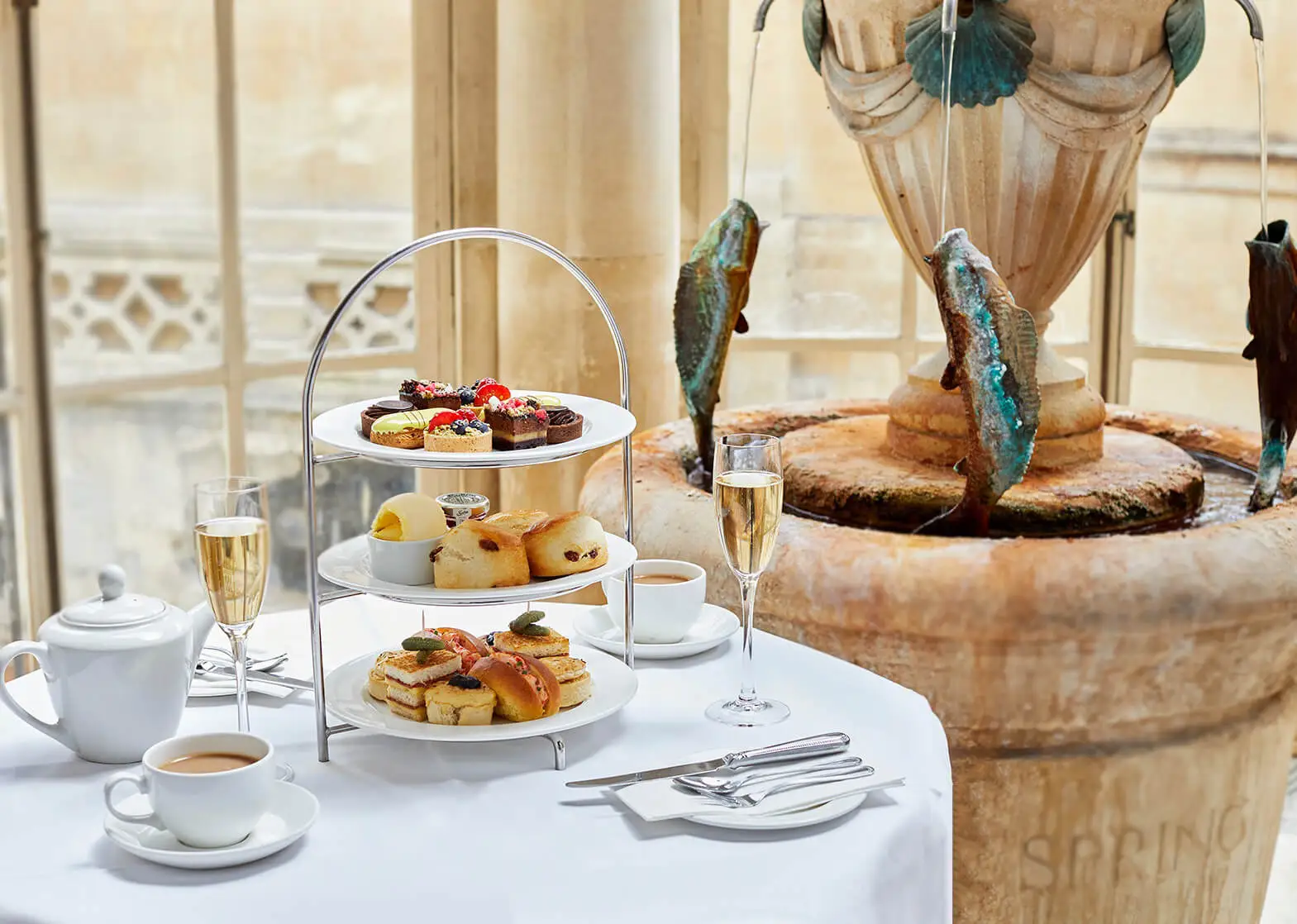 A formal table set for a traditional afternoon tea with a white table cloth, a three tiered tray of pastries, champagne and tea cups with a teapot inside the Pump Room at the Roman Baths in Bath, England. 