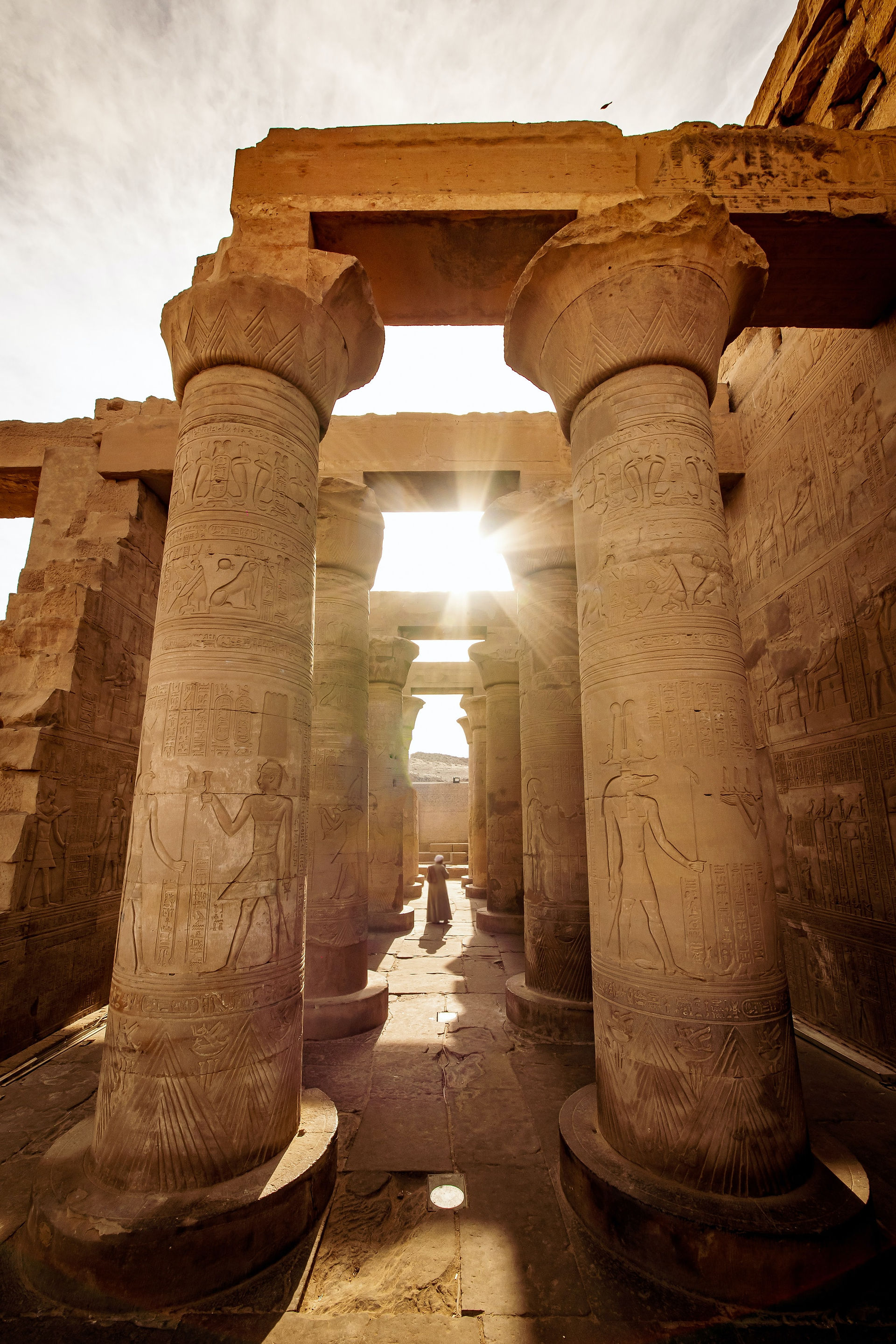 An ancient Egyptian temple called Kom Ombo with wide stone pillars, sand, and sunlight pouring through the sandstone sculptures and hieroglyphics.