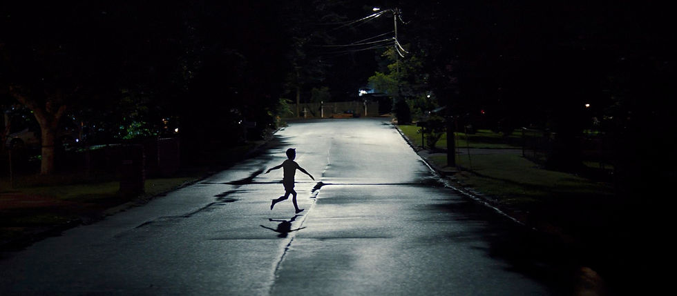 A child runs on a dimly lit street at night, surrounded by trees and a few scattered houses. The mood is mysterious and quiet.
