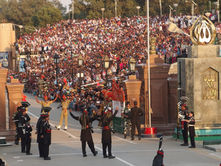 Border ceremony with uniformed guards performing, large crowd in stands cheering. Stone columns display inscriptions. Energetic atmosphere.